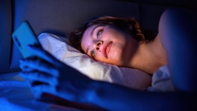 Mujer usando el móvil en la cama de noche, un hábito que dificulta conciliar el sueño